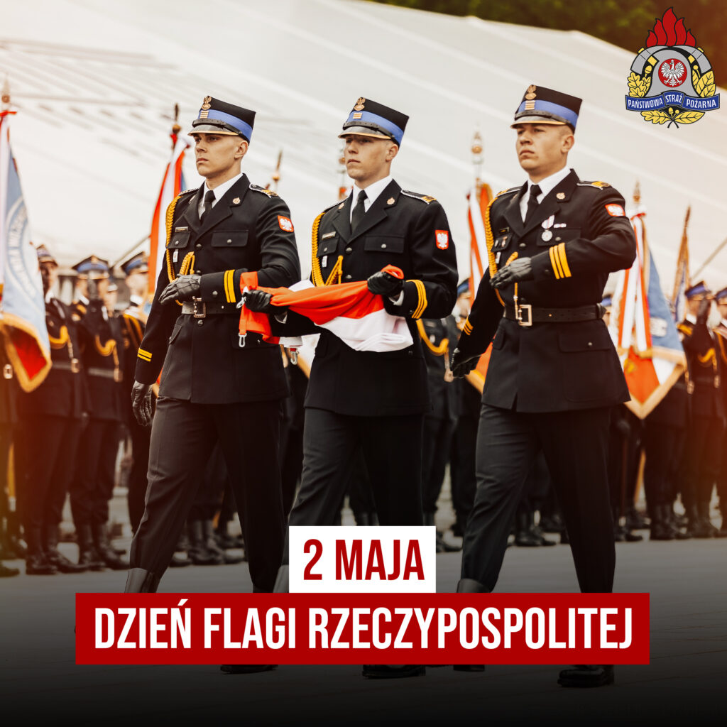 Trzech strażaków Państwowej Straży Pożarnej w mundurach galowych niesie złożoną flagę Polski podczas uroczystego przemarszu. W tle poczty sztandarowe. Napis: „2 maja – Dzień Flagi Rzeczypospolitej”.