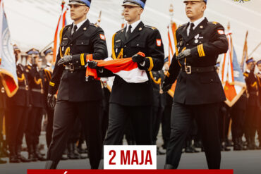 Trzech strażaków Państwowej Straży Pożarnej w mundurach galowych niesie złożoną flagę Polski podczas uroczystego przemarszu. W tle poczty sztandarowe. Napis: „2 maja – Dzień Flagi Rzeczypospolitej”.